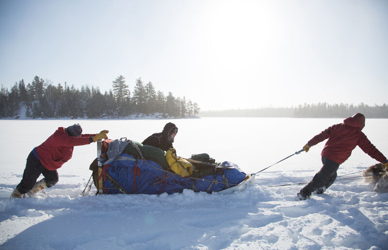 In a snowy landscape, three people are pulling a sled loaded with gear. Two individuals are pushing from behind, while another pulls from the front, assisted by a dog. They are traversing a snow-covered field, possibly a frozen lake, with a line of trees visible in the background. The sky is bright, suggesting a sunny day.